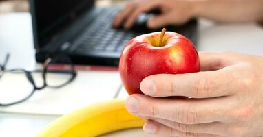 The concept of a healthy snack at work. A young man is holding an apple and working on a laptop at home or at work. Close-up. Selective focus. photo