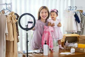 Young Asian influencer mother is selling children clothes live online streaming at home while raising her toddler as assistance photo