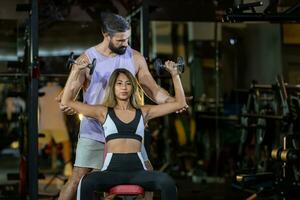 Asian woman is using dumbbell in weight training for arm muscle inside gym with support from trainer to prevent injury dark background for exercising and workout photo