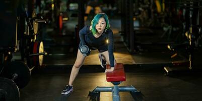 Asian woman with strong and muscular body training on bent over row dumbbell with bench staring at the camera to build back and wing muscle inside the gym with dark tone style photo