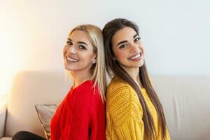 Two beautiful women with blond and brunette hair wearing knitted jumpers sitting on the sofa, back to back and smilling photo