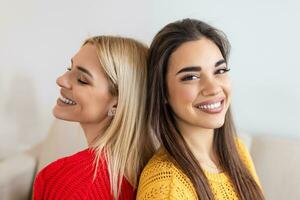 Two beautiful women with blond and brunette hair wearing knitted jumpers sitting on the sofa, back to back and smilling, Cheerful women back against back looking at camera and sitting on the sofa photo