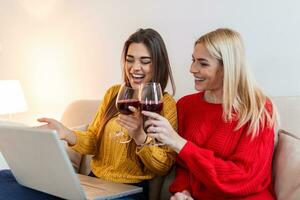 Two female friends spending time together at home, using laptop and digital tablet, sitting on sofa. copy space. Cheerful women websurfing, drinking wine photo