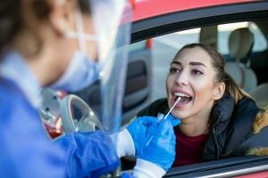 médico trabajador ejecutando Auto-servicio covid-19 comprobar, tomando nasal torunda muestra muestra desde hembra paciente mediante coche ventana, pcr diagnóstico para coronavirus presencia, doctor en ppe participación prueba equipo foto