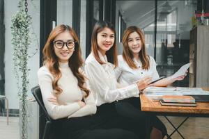 Three happy beautiful business woman diverse sitting on office desk and looking at camera, portrait photo