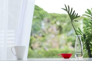 cup of coffee is prepared on table next to glass vase with leaves inserted to decorate table in beautiful modern style. cup of hot coffee on table by window in  morning and copy space for text. photo