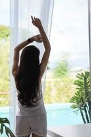 back silhouette of woman wearing pajamas stands by  window and writhes to relieve sleepiness after waking up in morning. girl woke up in morning alone and was twisting to ease her sleepiness. photo