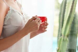 Young woman wearing white see through pajamas standing by window in morning and holding cup of coffee to drink and see beautiful morning scenery alone.  concept of drinking coffee to stimulate body. photo