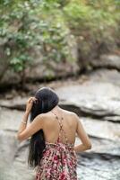 joven mujer turista en Clásico vestir soportes solo en rock por cascada en el bosque esperando a frio abajo para nadar. el espalda de joven asiático mujer turista quien disfruta jugando en cascada solo. foto