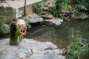 young female tourist in vintage dress sits alone on rock by waterfall in the forest hoping to cool down for swim. The back of young Asian female tourist who enjoys playing in waterfall alone. photo