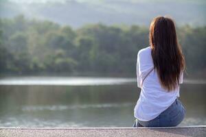 young woman sits alone on a path by a reservoir, lonely and depressed after living alone for so long. The back of a woman sitting by the reservoir alone in the evening and with a copy space for text. photo