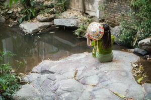 young female tourist in vintage dress sits alone on rock by waterfall in the forest hoping to cool down for swim. The back of young Asian female tourist who enjoys playing in waterfall alone. photo