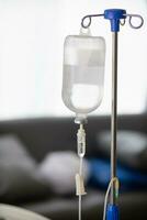 An iron stand to hang saline bottle high to deliver saline via catheter to an intravenous patient lying on patient bed. Medical concept in which doctor gives saline solution to patient through vein photo