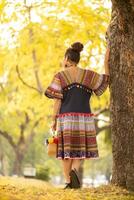 An elderly woman wearing fashionable dress using traditional tribal fabric designed to match era and standing under beautiful yellow flowering tree. An elderly woman stood alone under yellow flower. photo