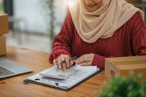 un hermosa y contento joven asiático musulmán hembra en línea vendedor utilizando un calculadora, comprobación productos cepo y calculador ventas y ingresos a su escritorio. foto