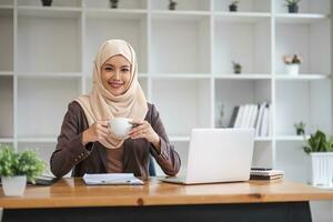 un retrato de un hermosa y exitoso asiático musulmán mujer de negocios o hembra jefe en hiyab, sonriente y mirando a el cámara mientras sentado a su escritorio con un café taza en su manos. foto