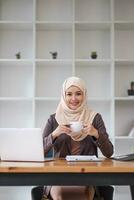 un retrato de un hermosa y exitoso asiático musulmán mujer de negocios o hembra jefe en hiyab, sonriente y mirando a el cámara mientras sentado a su escritorio con un café taza en su manos. foto