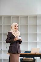 A portrait of a confident and smiling Asian Muslim businesswoman stands in her modern office with a coffee cup in her hands. photo