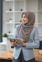 un retrato de un exitoso asiático musulmán mujer de negocios soportes en su moderno oficina con un digital tableta en su mano. foto