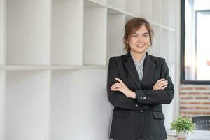 retrato, joven confidente sonriente asiático negocio mujer vistiendo traje en pie en oficina con brazos cruzado. foto