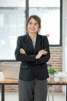 retrato, joven confidente sonriente asiático negocio mujer vistiendo traje en pie en oficina con brazos cruzado. foto