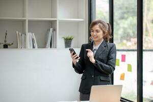 Asian businesswoman talking on mobile phone working on laptop in modern office. Happy business woman talking on mobile phone while analyzing weekly schedule in laptop photo