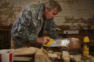 Mature carpenter sawing wood with a saw in his workshop for making wooden toys photo