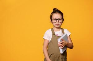 Studio portrait of Caucasian confident serious charming little girl 6 years old, holding textbook over orange background photo