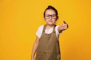 Serious pupil girl in teacher's glasses, points finger at camera, showing warning hand sign, isolated orange background photo