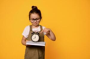 adorable niño, pequeño Chica de escuela en los anteojos, señalando su índice dedo a alarma reloj. es hora a espalda a escuela. foto