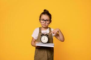 Cute primary school girl in eyeglasses, holding a book and showing a black alarm clock and looking confidently at camera photo