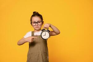 Adorable elementary age child points at a black alarm clock, shows the time to go to school, isolated orange background photo