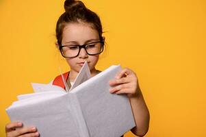 Close-up school girl carries school bag, reads book, standing isolated on orange background. Kids and education concept photo