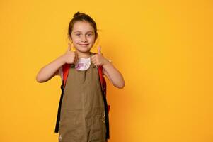 Adorable primary school girl carrying a backpack, smiles looking at camera, thumbs up, expressing positive emotions photo