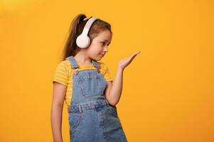 Cute little child girl wearing wireless headphones, smiles looking at her hand palm up, isolated orange studio backdrop photo