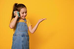 sonriente contento pequeño niña en azul mezclilla vestido, negociaciones en inteligente móvil teléfono, mira a imaginario Copiar espacio en su mano foto