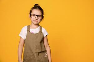 Mischievous child girl in stylish casual clothes and eyeglasses, looking at camera, isolated orange studio background photo