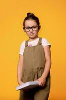 Caucasian cute girl in eyeglasses, smiles looking at camera, holds a textbook isolated over orange studio background photo