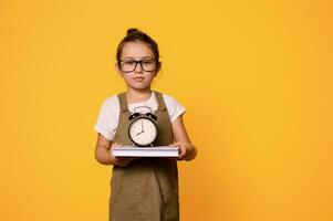 Authentic primary school student in eyeglasses, holds a book and alarm clock, isolated orange background. Copy ad space photo