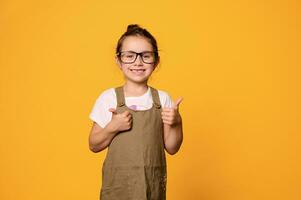 Happy little girl in eyeglasses, gestures with thumbs up, isolated orange studio background. Smart pupil. Teacher's Day photo