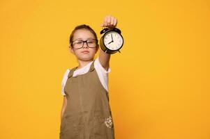 A black alarm clock in girl's hand, showing time on the dial, isolated orange backdrop. Back to school. Time management photo
