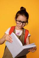 primario colegio niña en casual vestir y anteojos, aprende leyendo alfabeto, participación Bosquejo libro, aislado naranja antecedentes foto