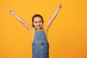 Happy smiling cheerful preschool girl, clenching fists and raising arms, expressing positive emotions isolated on orange photo