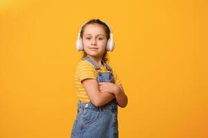 Cute little child girl with arms folded, in wireless headphones, smiles looking at camera, isolated on orange background photo