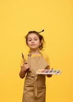 Inspired creative talented kid girl holding watercolor palette, paint brush and palette knife, isolated yellow backddrop photo