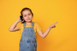Charming kid girl 6 years old, talking on mobile phone, pointing finger aside copy ad space, isolated yellow background photo
