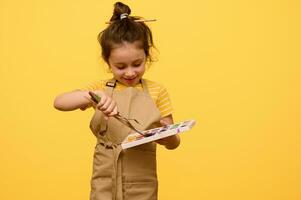 sonriente pequeño niña artista inmersión Cepillo de pintura dentro acuarela pinturas, aislado amarillo antecedentes. Copiar publicidad espacio foto