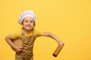 Kid girl in chef hat and apron, makes faces, looking at camera, holding rolling pin and wooden board, isolated on yellow photo
