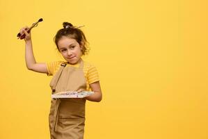 Creative child, little girl painter in apron with paintbrush in hairstyle, holds watercolor palette and painting tools photo