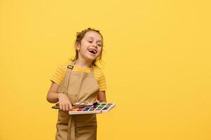 Caucasian child girl laughing, expressing positive emotions during art class, holding watercolor paints and paintbrush photo